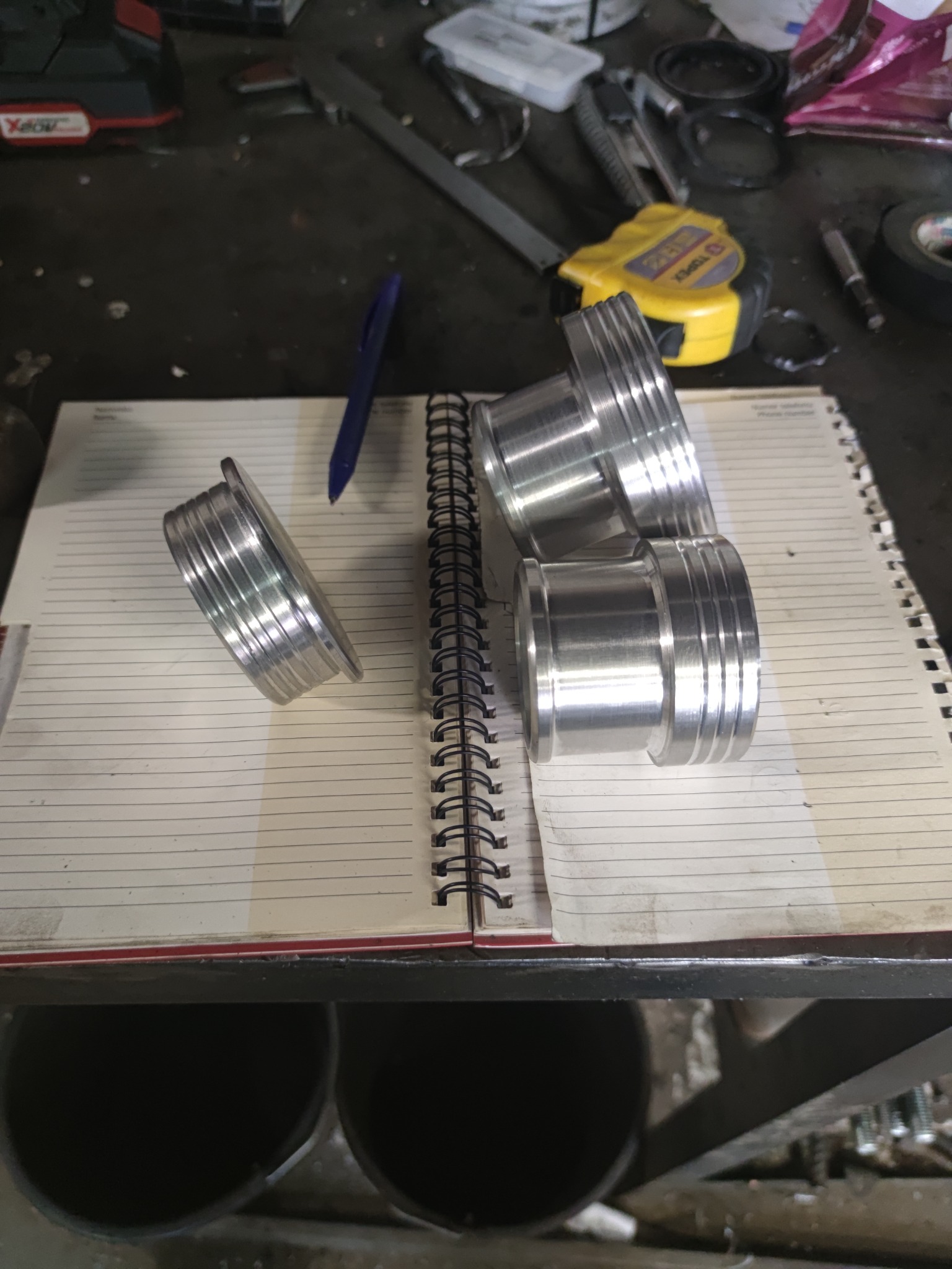 Cztery precyzyjnie obrobione, aluminiowe elementy z rowkami, leżące na otwartym notesie w warsztacie, obok długopisu i spiralnego grzbietu notesu; w tle widoczne narzędzia i miarka.