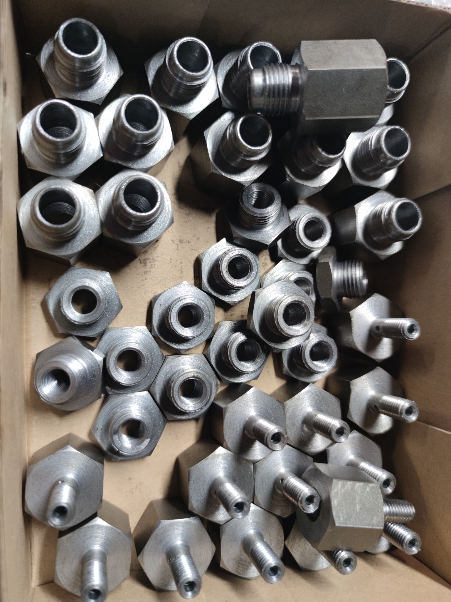 Srebrne, metalowe złączki hydrauliczne o heksagonalnej podstawie i gwintowanych otworach, ułożone w kartonowym pudełku, widoczne ślady obróbki skrawaniem.