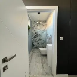 Wnętrze łazienki z motywem kwiatowym na ścianie, widok z korytarza przez otwarte białe drzwi z ciemnym uchwytem, wisząca szklana lampa, umywalka na postumencie i toaleta.