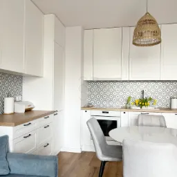 Jasna kuchnia z białymi szafkami, drewnianym blatem i wzorzystymi płytkami backsplash, uzupełniona okrągłym stołem i szarymi krzesłami oraz wiklinową lampą.