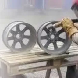 Pracownik w kombinezonie ochronnym czyści strumieniowo felgę samochodową na drewnianej palecie, obok stoi druga felga.