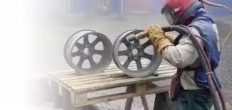 Pracownik w kombinezonie ochronnym czyści strumieniowo felgę samochodową na drewnianej palecie, obok stoi druga felga.