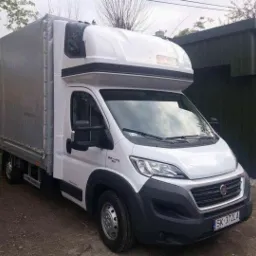 Biały samochód dostawczy z plandeką w Mysłowicach, zaparkowany przed zielonym budynkiem gospodarczym.