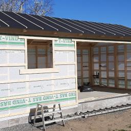 Bygg service - Surowa konstrukcja altany z drewnianą ramą, folią wiatroizolacyjną i częściowo pokrytym dachem, widziana w słoneczny dzień z drabiną opartą o ścianę.