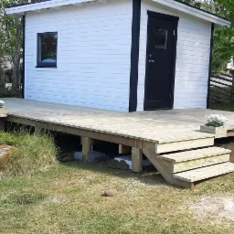 Nowo wybudowany drewniany taras z schodami, przylegający do małego białego domku z czarnymi detalami, na trawiastym terenie z naturalnymi kamieniami i roślinnością.