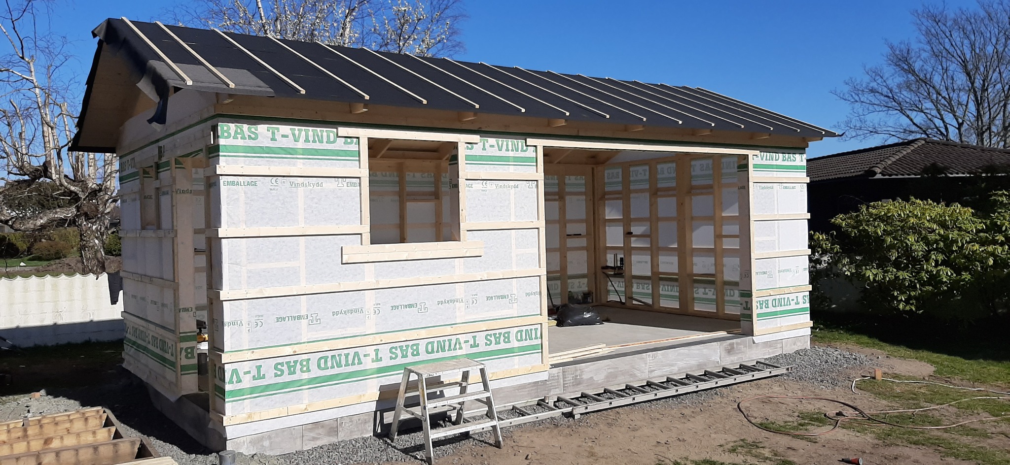 Surowa konstrukcja altany z drewnianą ramą, folią wiatroizolacyjną i częściowo pokrytym dachem, widziana w słoneczny dzień z drabiną opartą o ścianę.