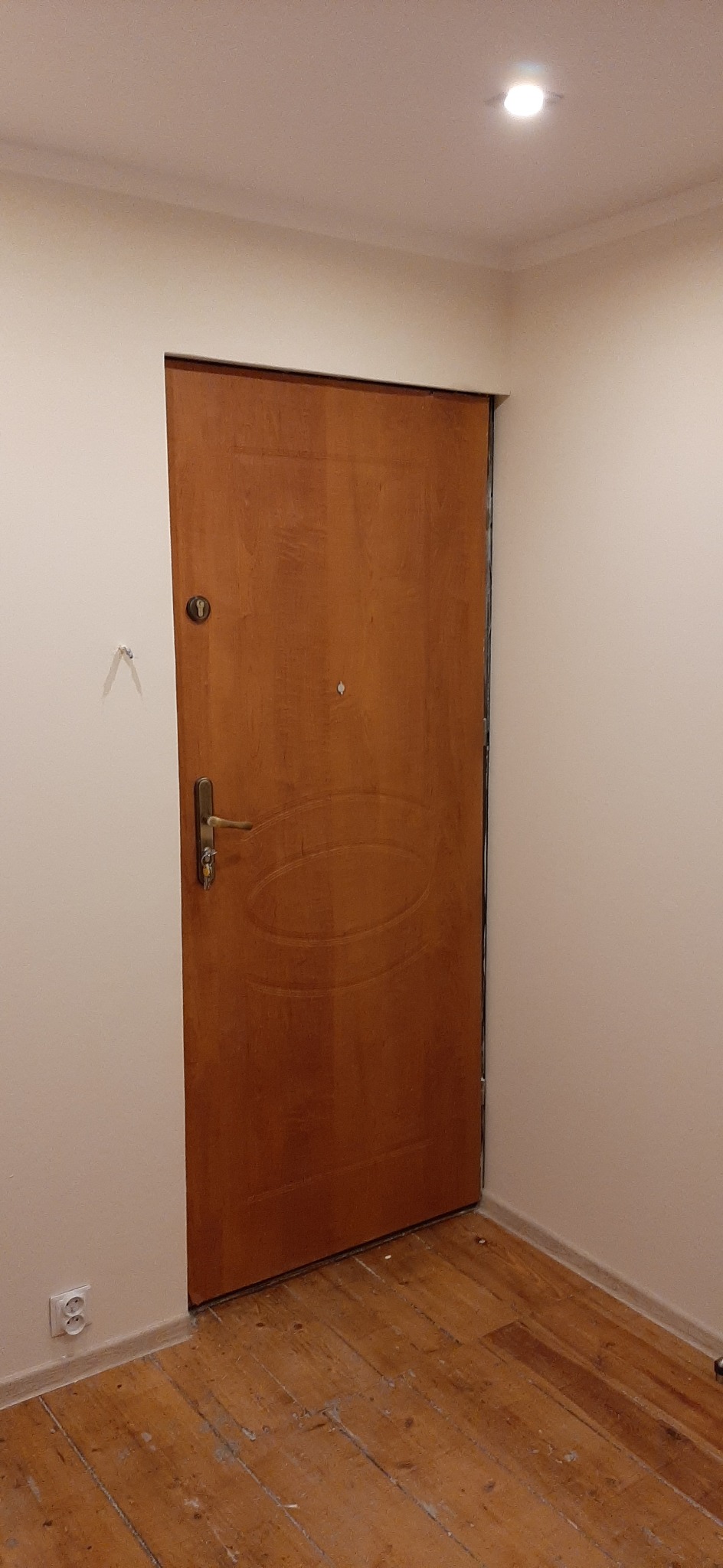 Brązowe drzwi wejściowe z widoczną klamką i zamkiem, osadzone w ościeżnicy w jasnym korytarzu z drewnianą podłogą, widoczne ślady po niedokończonym montażu.