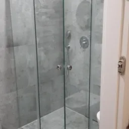 Szklana kabina prysznicowa narożna z deszczownicą i baterią, zamontowana w łazience wykończonej szarymi płytkami imitującymi kamień.