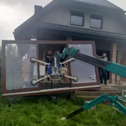 Montaż dużego okna z brązową ramą za pomocą specjalistycznego dźwigu z przyssawkami na tle domu w trakcie budowy.