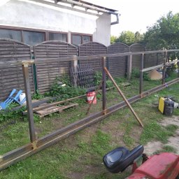 Ad- Mat - Metalowa konstrukcja ogrodzenia w trakcie budowy na trawniku, z widocznym sprzętem spawalniczym i elementami drewnianego płotu w tle.