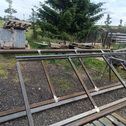 Ad- Mat - Metalowe ramy konstrukcyjne ze spawami, leżące na ziemi, w tle przyczepa z paletami i drzewa. Widoczne elementy konstrukcji stalowych przed montażem.