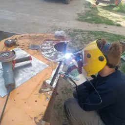 Spawanie elementu na zewnątrz: spawacz w masce łączy metalowe części na pomarańczowym stole roboczym, obok szlifierka kątowa. W tle trawa i zaparkowane samochody.