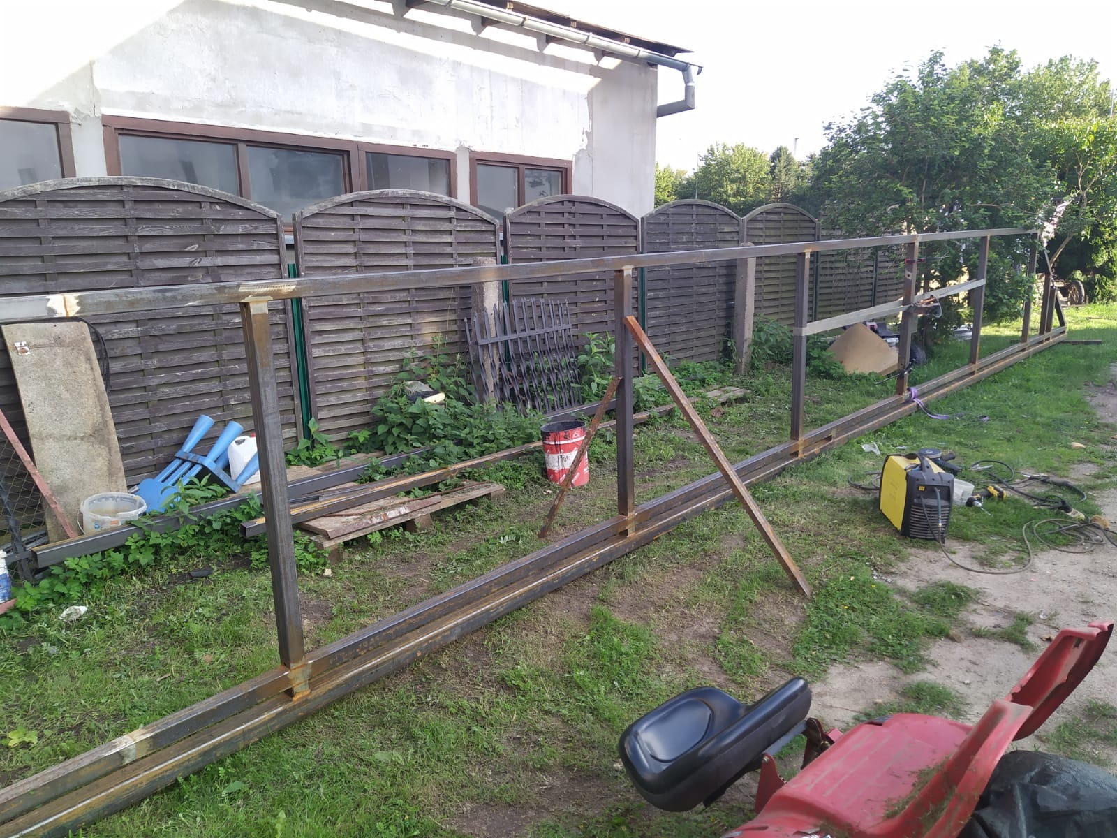 Metalowa konstrukcja ogrodzenia w trakcie budowy na trawniku, z widocznym sprzętem spawalniczym i elementami drewnianego płotu w tle.