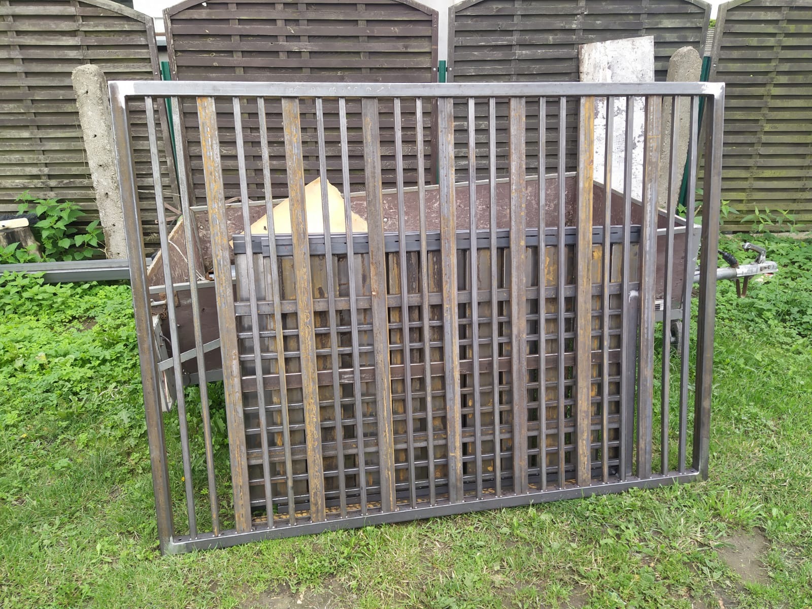 Surowa, metalowa konstrukcja bramy wjazdowej z prostokątnymi kratami, leżąca na trawie przed drewnianym płotem. Widoczny fragment przyczepy.