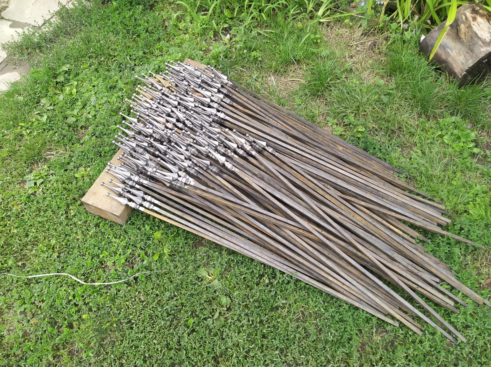 Wiele metalowych prętów z ozdobnymi grotami, ułożonych na trawie i drewnianym klocku. Elementy do balustrady lub bramy wjazdowej, przygotowane do montażu.