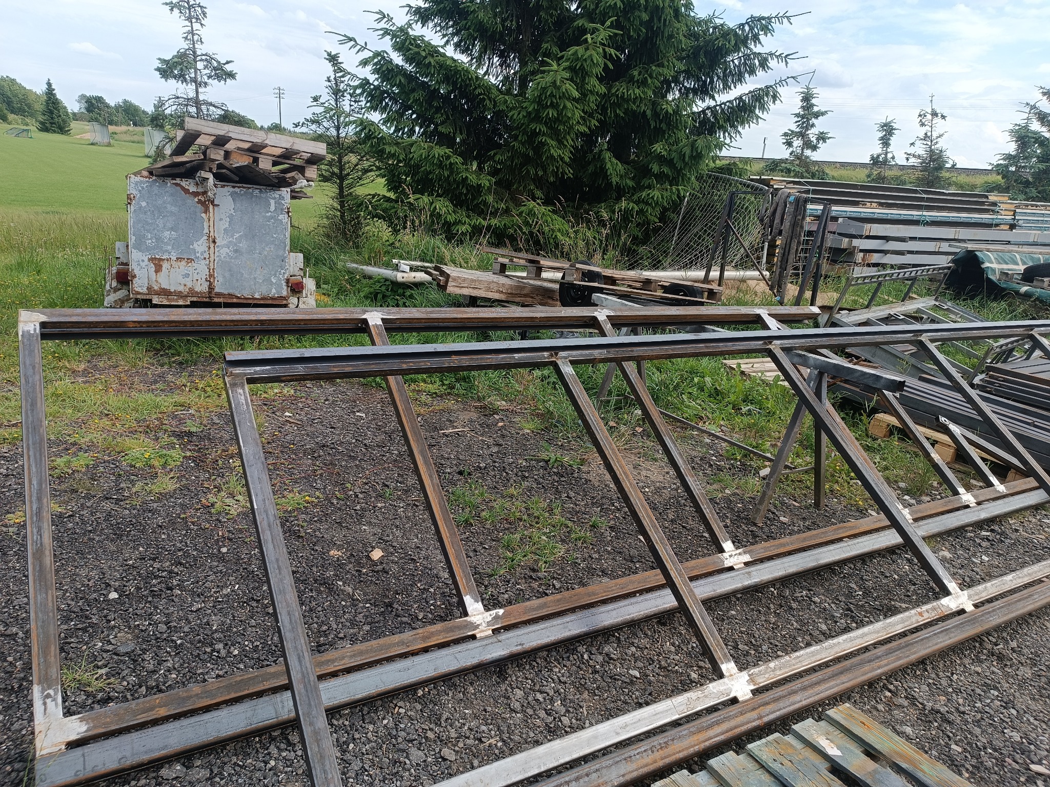 Metalowe ramy konstrukcyjne ze spawami, leżące na ziemi, w tle przyczepa z paletami i drzewa. Widoczne elementy konstrukcji stalowych przed montażem.