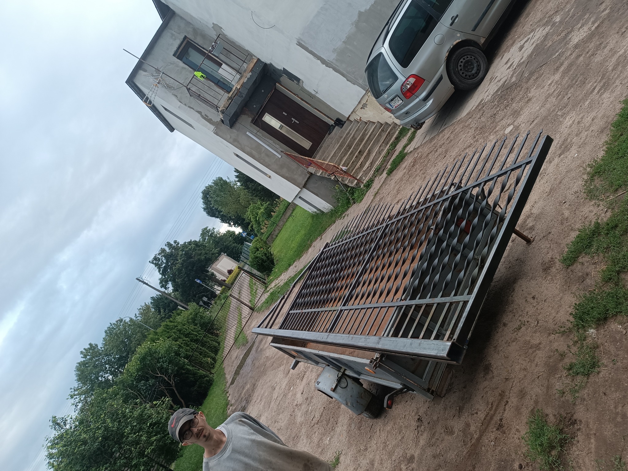 Brama wjazdowa z metalowymi zdobieniami na przyczepce w Chojnicach, gotowa do montażu. W tle dom z balkonem i samochód.