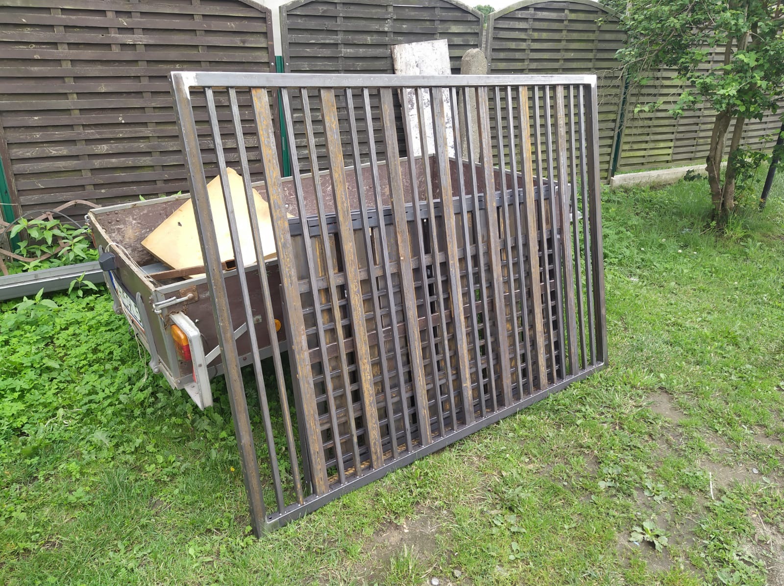 Surowa, metalowa konstrukcja kratowa, prawdopodobnie fragment bramy, oparta o przyczepkę na trawie, na tle drewnianego płotu i drzewa. Widoczne ślady spawania.