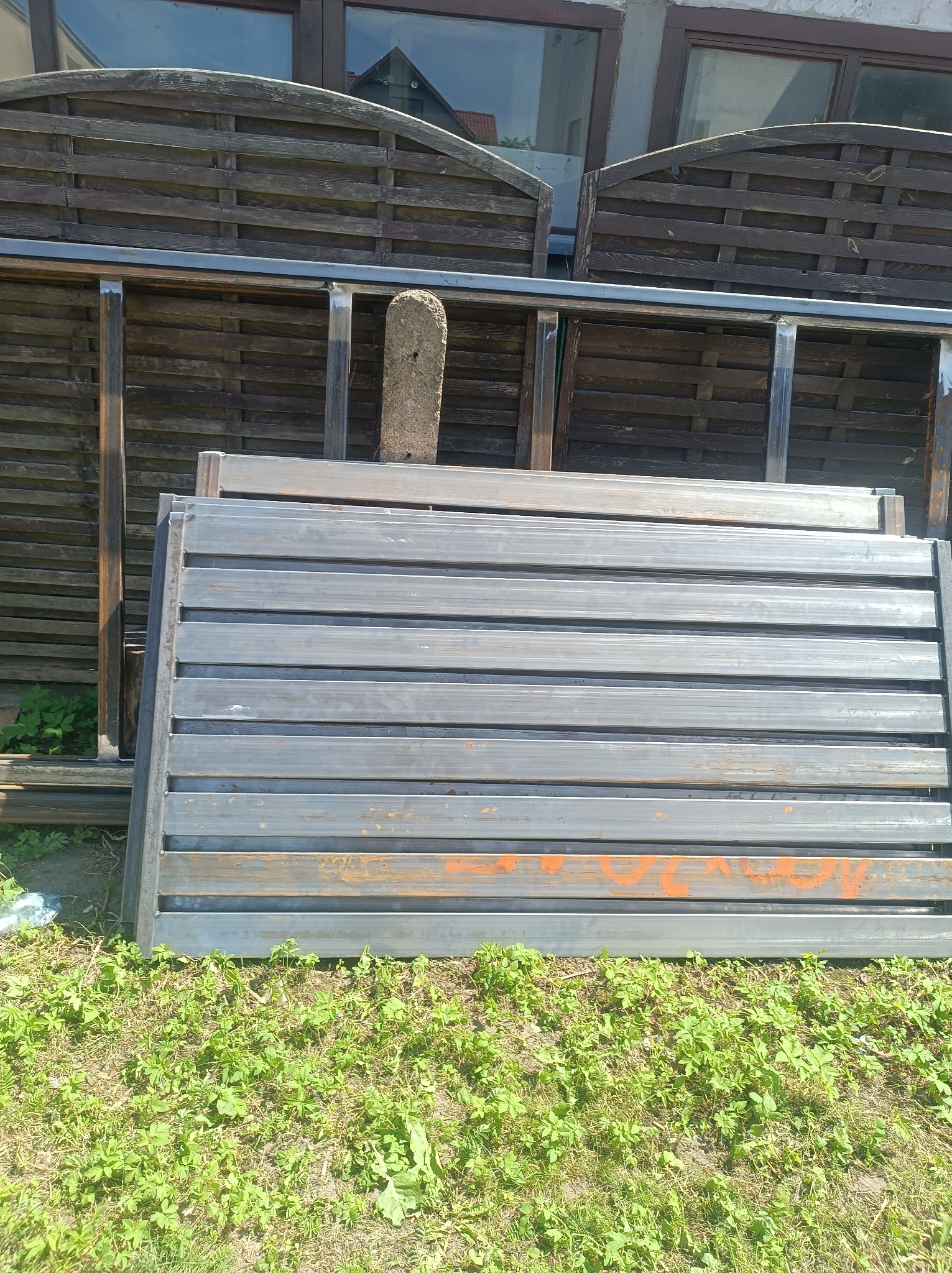 Stos elementów bramy wjazdowej z metalowych profili, leżący na trawie, z drewnianym płotem w tle. Widoczne ślady pomarańczowej farby na jednym z elementów.