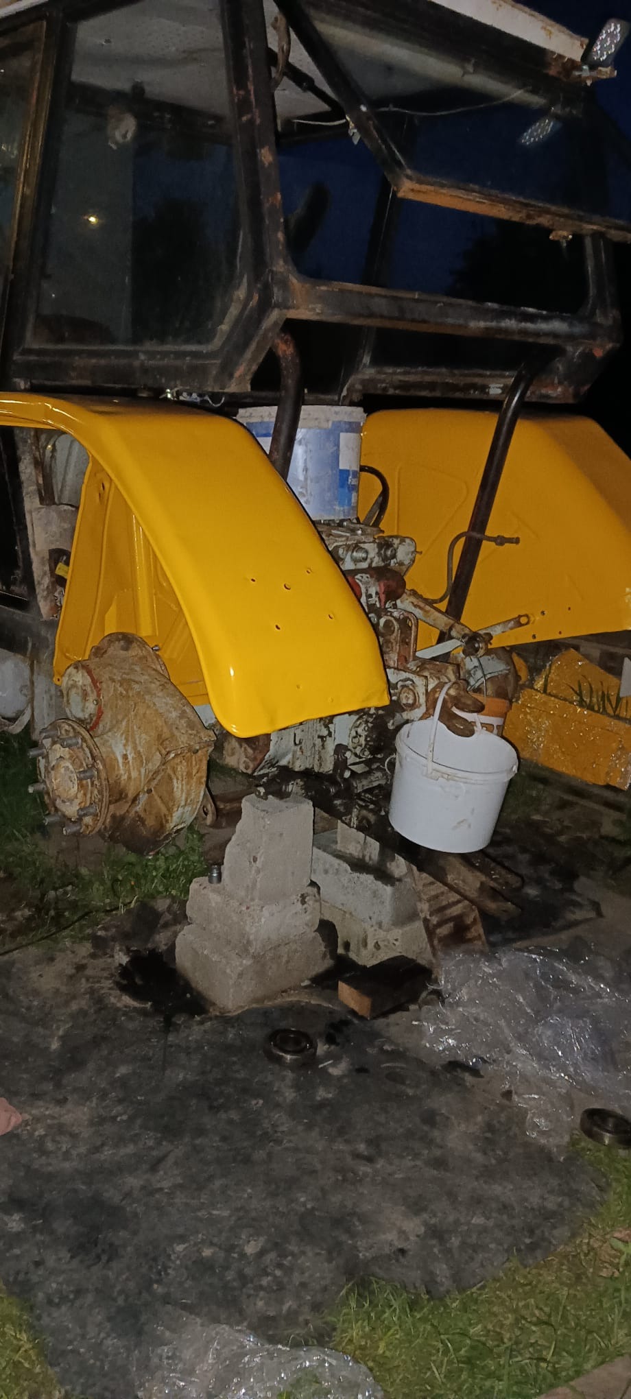 Fragment odrestaurowanego traktora z żółtymi błotnikami i elementami podwozia, widoczny w słabym oświetleniu, sugerujący etap prac warsztatowych.