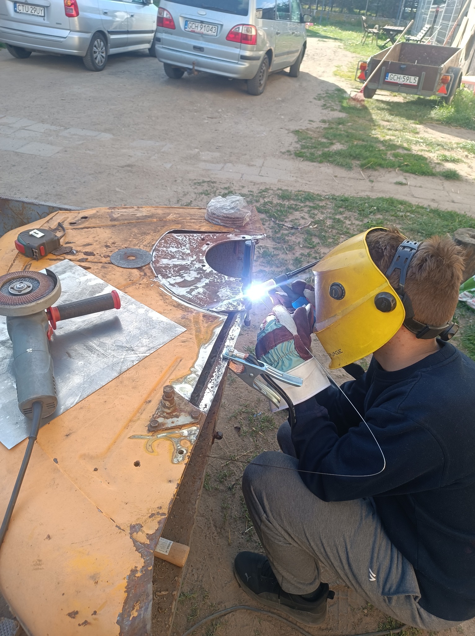 Spawanie elementu na zewnątrz: spawacz w masce łączy metalowe części na pomarańczowym stole roboczym, obok szlifierka kątowa. W tle trawa i zaparkowane samochody.