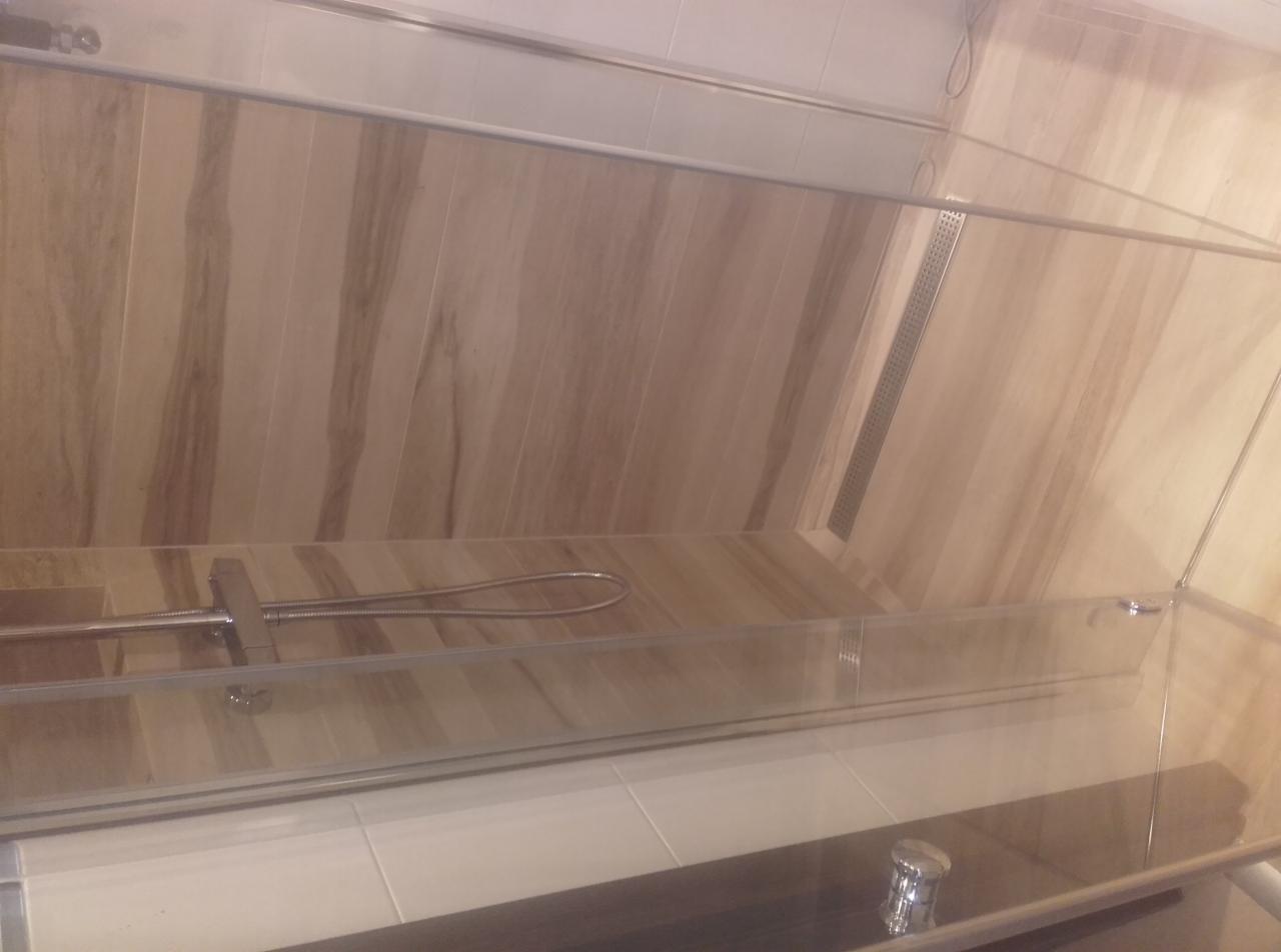 Kabina prysznicowa z odpływem liniowym w posadzce, wykończona płytkami imitującymi marmur w odcieniach beżu i brązu, widoczna bateria prysznicowa.