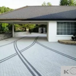 Podjazd do domu wyłożony jasną kostką z ciemnymi pasami tworzącymi łukowe wzory, widoczny zadaszony wjazd i fragment elewacji budynku.