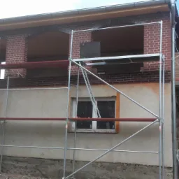 Dom w trakcie prac elewacyjnych z widocznymi rusztowaniami, fragmentami tynku i ceglanymi elementami dekoracyjnymi nad oknami.