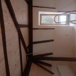 Poddasze z drewnianą konstrukcją, częściowo ocieplone i wykończone płytami, widoczny fragment drabiny malarskiej i wiadro z farbą.