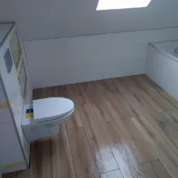 Wykończona łazienka na poddaszu z drewnopodobną podłogą, białą armaturą i oknem dachowym.