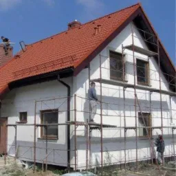Dom jednorodzinny w trakcie ocieplania ścian zewnętrznych, widoczne rusztowania, styropianowe płyty i pracownicy w trakcie montażu. Czerwony dach kontrastuje z białą elewacją.