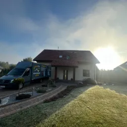 Szary samochód dostawczy z reklamą wełny wdmuchiwanej zaparkowany przed domem jednorodzinnym w słoneczny poranek.