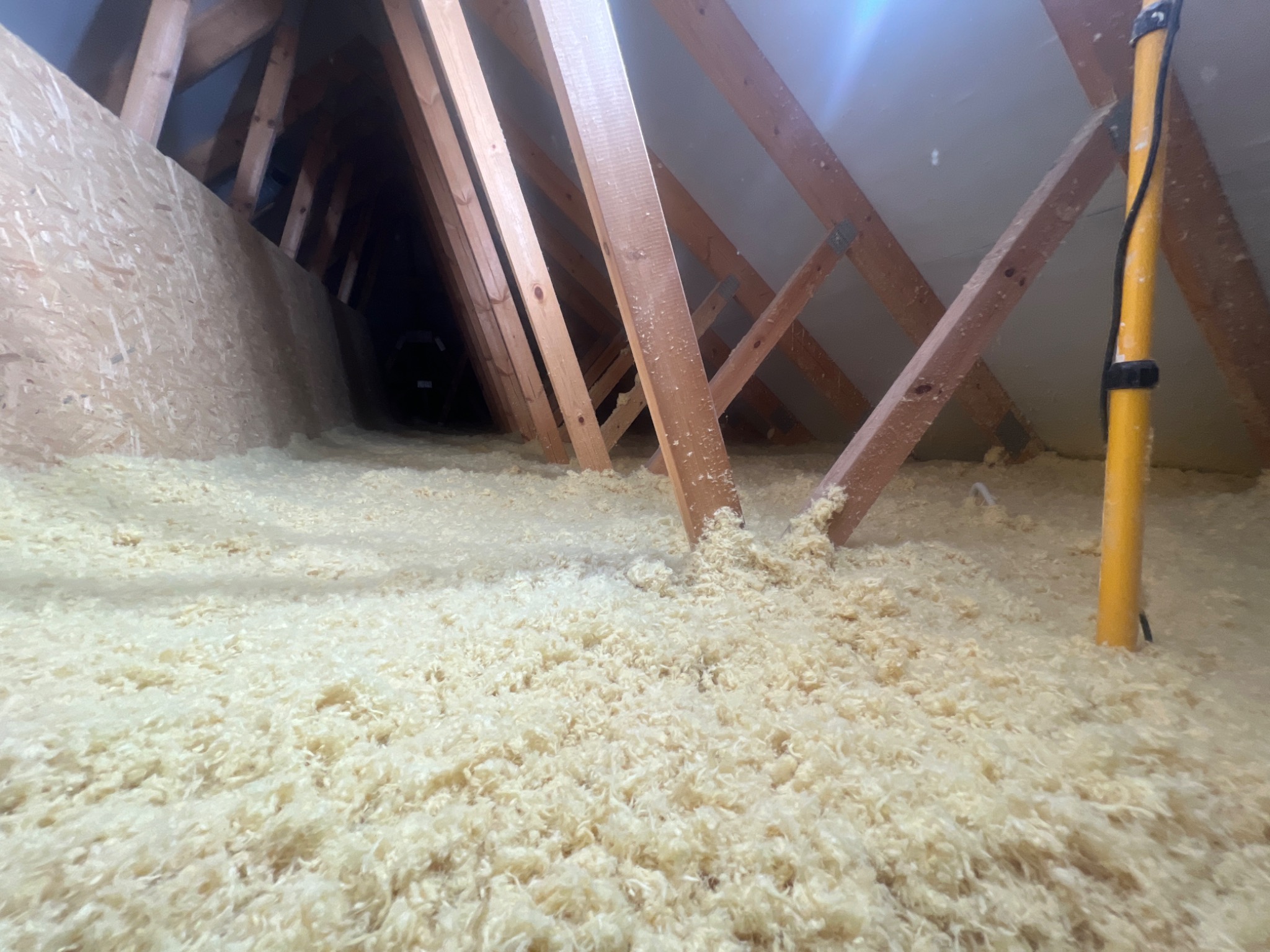 Poddasze z widoczną drewnianą konstrukcją dachu i warstwą izolacji z wełny celulozowej, oświetlane lampą roboczą na żółtym statywie.