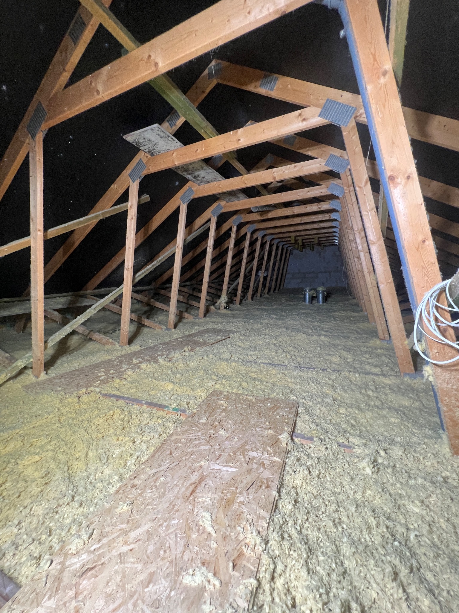 Poddasze z drewnianą konstrukcją więźby dachowej, częściowo ocieplone wełną mineralną, z widocznymi fragmentami płyt OSB na podłodze i przewodami elektrycznymi.