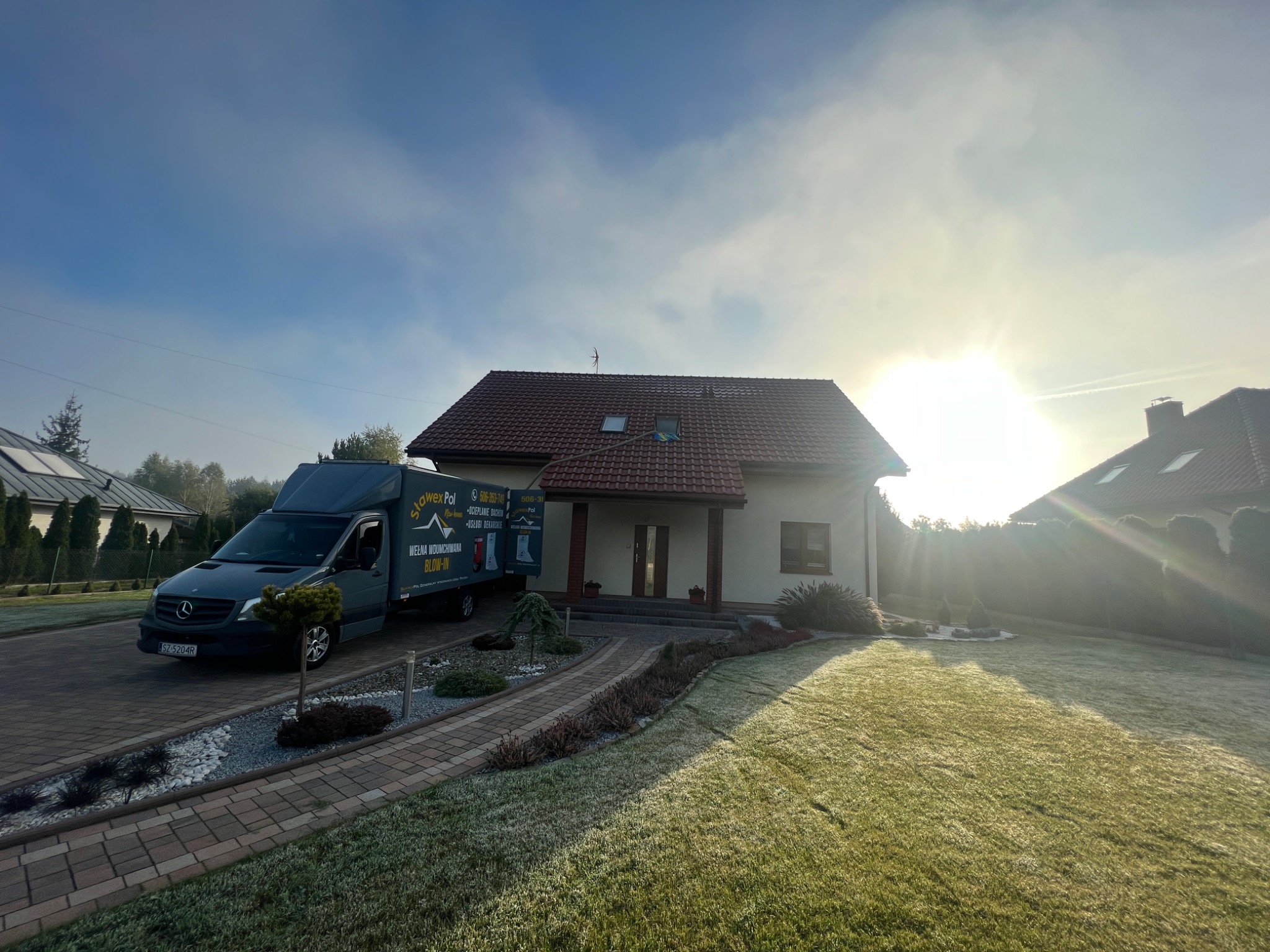 Szary samochód dostawczy z reklamą wełny wdmuchiwanej zaparkowany przed domem jednorodzinnym w słoneczny poranek.