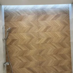 Wykończona ściana prysznicowa z płytek imitujących drewno ułożonych w jodełkę, z widoczną słuchawką prysznicową i baterią, oświetlona punktowo.