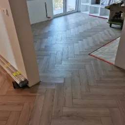 Ułożona podłoga z paneli w jodełkę, widoczne narzędzia i materiały budowlane, częściowo zabezpieczona folią ochronną.