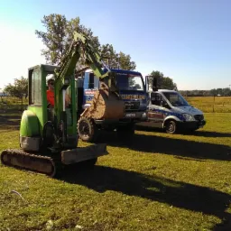 Zielona mini koparka na trawiastym terenie, obok zaparkowany samochód ciężarowy z napisem 'CENTESTUDNI' i bus Mercedes Sprinter z logiem firmy, w tle drzewa i błękitne niebo.