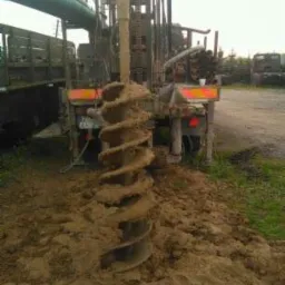 Wiertnica z świdrem spiralnym zagłębiającym się w ziemię, widoczne rury na platformie wiertniczej i otoczenie placu budowy.