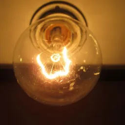 Świecąca żarówka o okrągłym kształcie, z widocznymi drobnymi zabrudzeniami na szkle, zawieszona pod sufitem, dająca ciepłe, żółte światło.