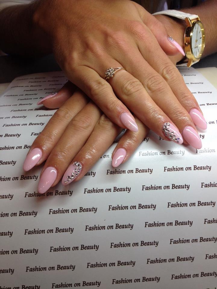 Dłonie kobiety z jasnoróżowym manicure hybrydowym, zdobionym cyrkoniami na dwóch paznokciach, na tle papieru z powtarzającym się napisem 'Fashion on Beauty'; na palcu serdecznym pierścionek...