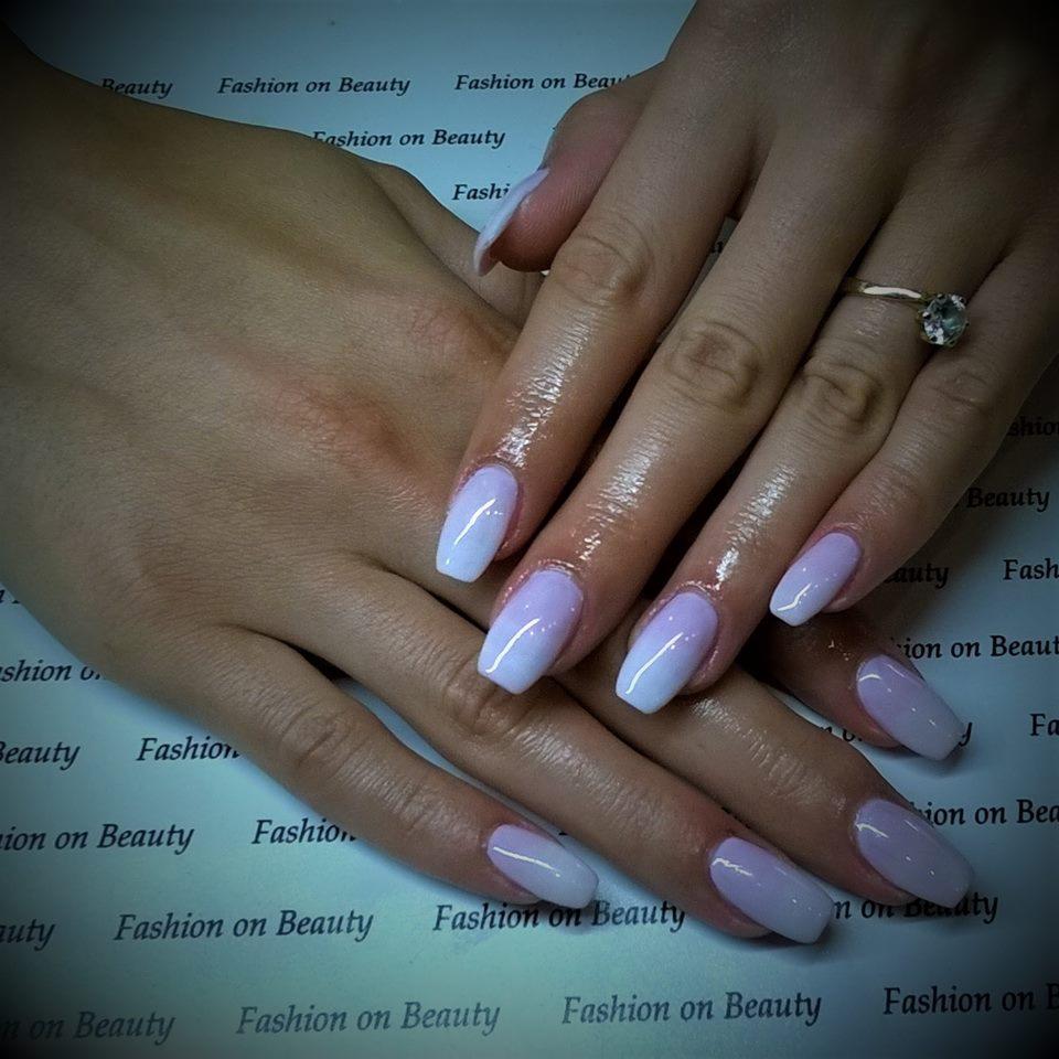 Dłonie z świeżo wykonanym manicure hybrydowym w odcieniach bieli i fioletu, kształt paznokci migdał, pierścionek z oczkiem na palcu serdecznym, tło z powtarzającym się napisem 'Fashion on Beauty'.