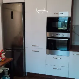 Biała zabudowa kuchenna z piekarnikiem, mikrofalówką i lodówką LG, uzupełniona o ciemną podłogę imitującą drewno.