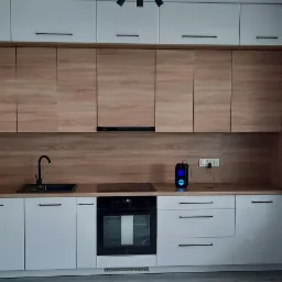 Nowoczesna zabudowa kuchenna z białymi szafkami dolnymi i górnymi w odcieniu drewna, czarny zlew i płyta grzewcza, widok na balkon zza szklanych drzwi.