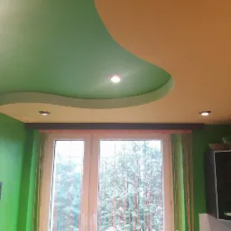 Pomarańczowo-zielone malowanie sufitu z dekoracyjną listwą i oświetleniem punktowym, widok na okno z zielonymi zasłonami i fragment zielonej ściany z obrazem.