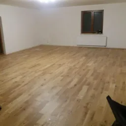 Jasna, drewniana podłoga ułożona w przestronnym pokoju z białymi ścianami, widoczne okno z brązową ramą, grzejnik pod oknem oraz czarny worek z narzędziami w rogu pokoju.