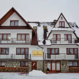 Dwupiętrowy budynek willi w stylu g&oacute;ralskim z białymi ścianami, brązowym dachem pokrytym śniegiem, kamienną podmur&oacute;wką i balkonami z czarnymi balustradami. Na ścianie szyld z napisem Willa Slavita.