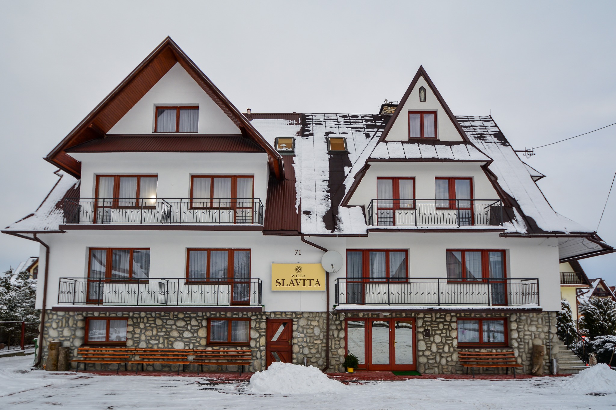 Dwupiętrowy budynek willi w stylu góralskim z białymi ścianami, brązowym dachem pokrytym śniegiem, kamienną podmurówką i balkonami z czarnymi balustradami. Na ścianie szyld z napisem Willa Slavita.