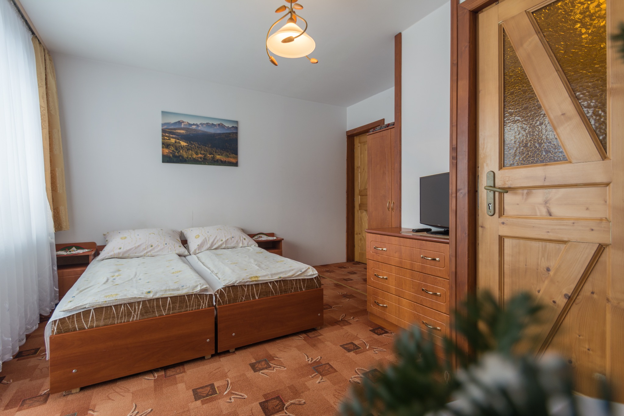 Wnętrze pokoju z dwoma łóżkami, drewnianymi meblami i obrazem gór na ścianie, widok na drewniane drzwi z szybą o fakturze szkła.