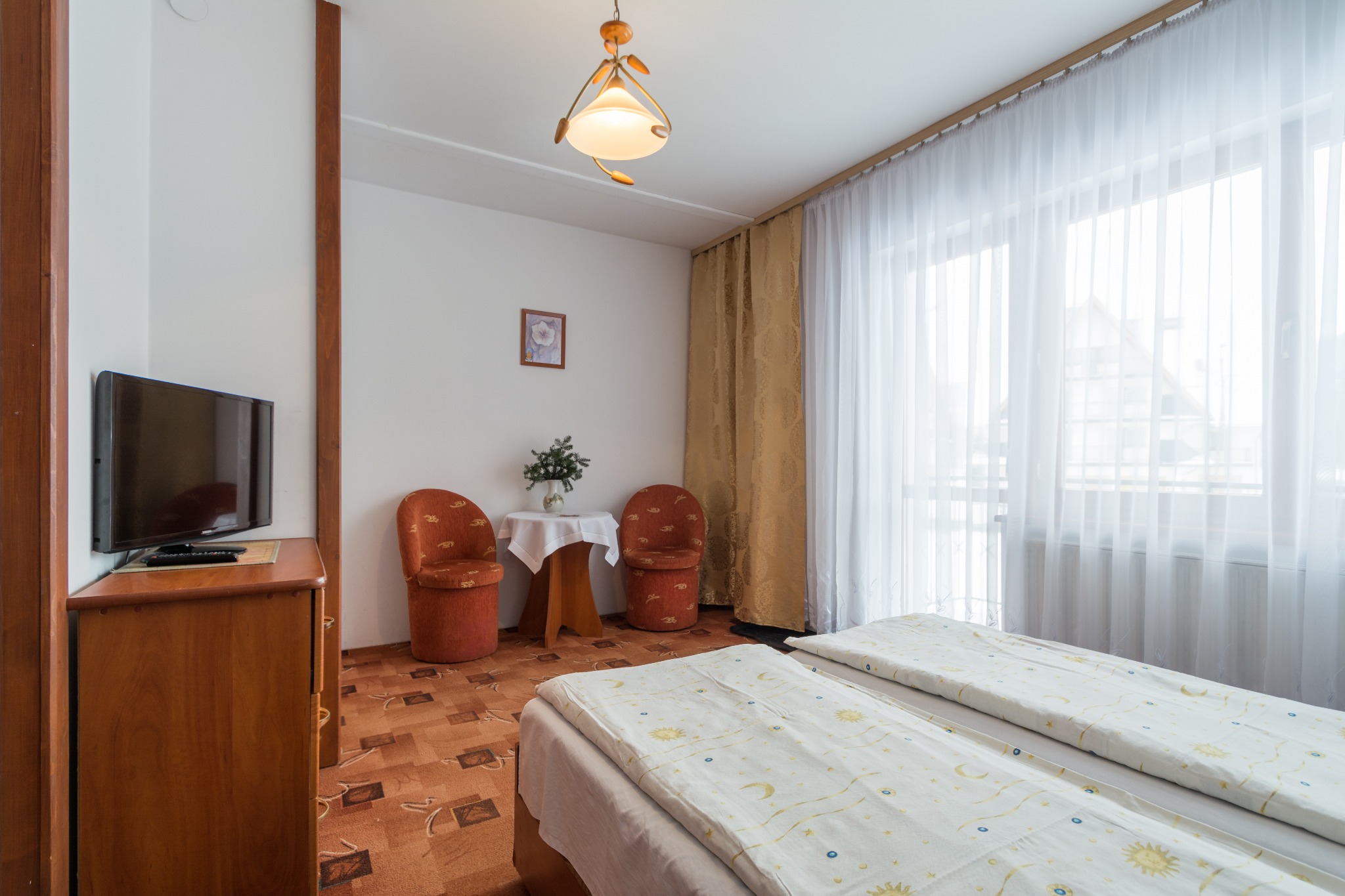 Jasne wnętrze pokoju z dwoma łóżkami, stolikiem i telewizorem, widok na okno z białymi zasłonami.